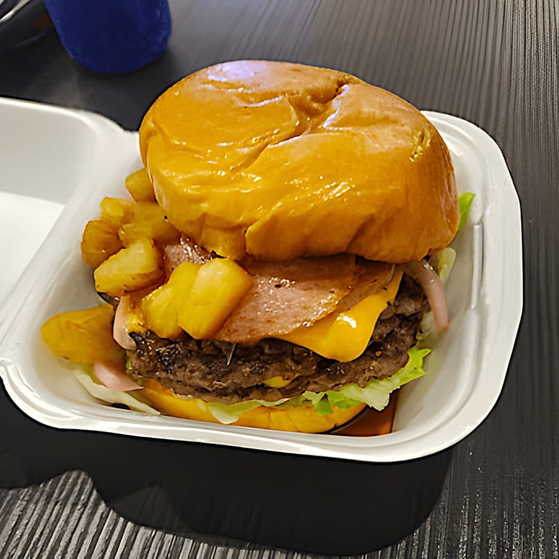 A double cheeseburger with pineapple, bacon, lettuce, and red onion in a white takeout container.