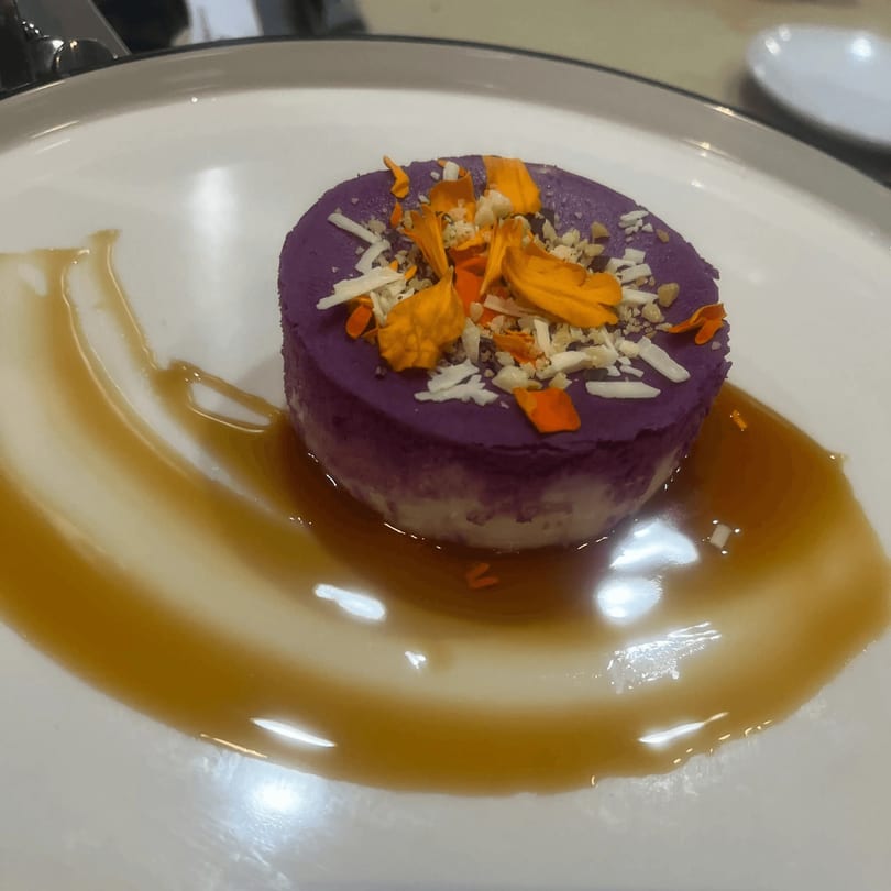 Purple ube dessert with orange flower petals, shredded coconut, and nuts, drizzled with caramel sauce.